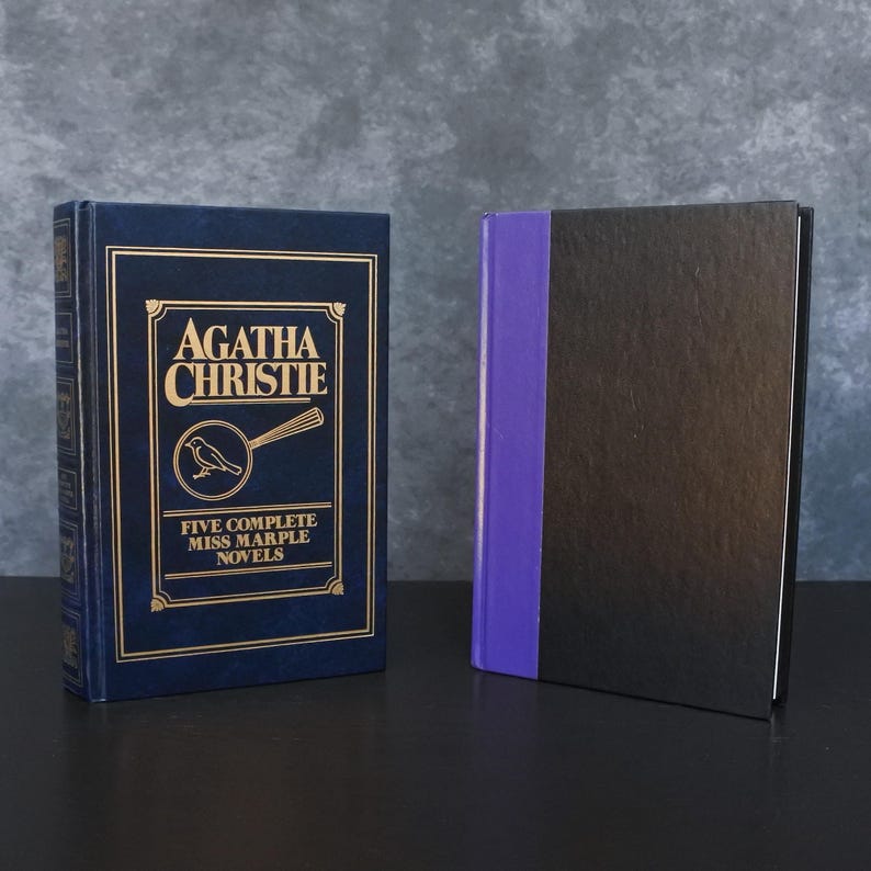 May include: Two hardcover books. The book on the left is dark blue with gold lettering that reads "Agatha Christie" and "Five Complete Miss Marple Novels." The book on the right is black with a purple spine.