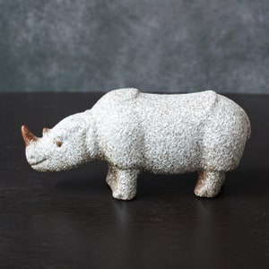 Op de afbeelding: Witte keramische neushoornfiguur met een getextureerd oppervlak. Het beeldje heeft een bruine hoorn en staat op een donker oppervlak.