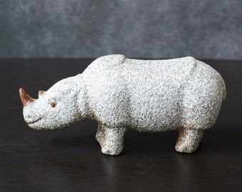 Op de afbeelding: Witte keramische neushoornfiguur met een getextureerd oppervlak. Het beeldje heeft een bruine hoorn en staat op een donker oppervlak.