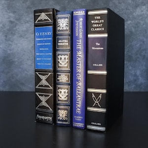 May include: A collection of hardcover books with decorative covers. Titles include works by O. Henry, Agatha Christie, and Robert Louis Stevenson. The books are in various colors, including black, blue, and gold, and are arranged vertically.