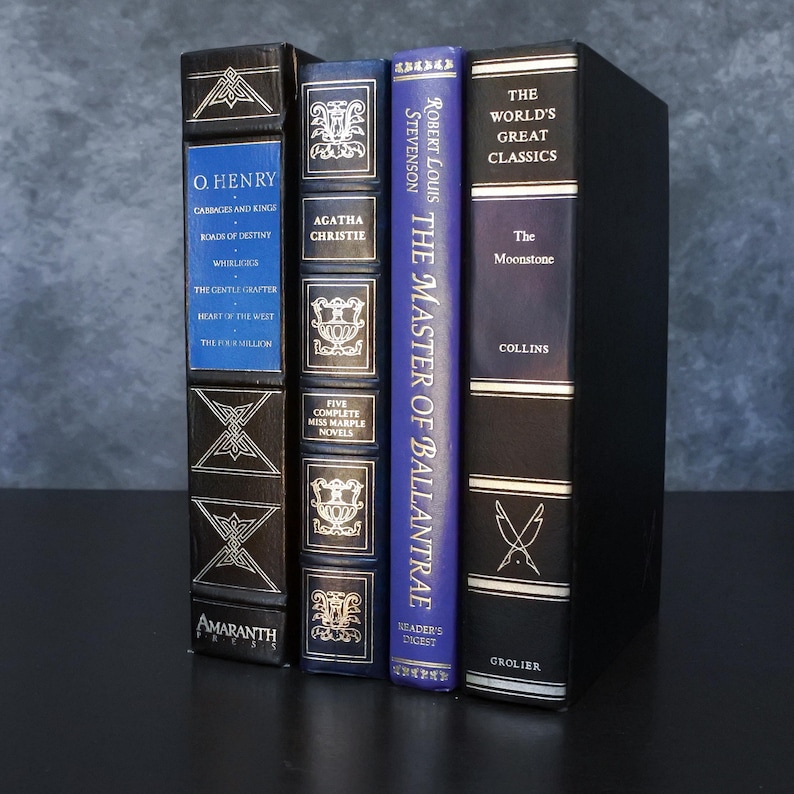 May include: A collection of hardcover books with decorative covers. Titles include works by O. Henry, Agatha Christie, and Robert Louis Stevenson. The books are in various colors, including black, blue, and gold, and are arranged vertically.