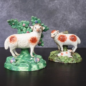 Puede incluir: Dos figuras de ovejas de cerámica. Una oveja está de pie cerca de un árbol verde con flores rosas y azules. La otra oveja está de pie con un cordero. Ambas ovejas tienen cuerpos blancos con manchas rojas y están sobre bases verdes.