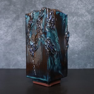 Könnte beinhalten: Eine rechteckige Keramikvase mit dunkelbrauner und türkisblauer Glasur. Die Vase hat strukturierte, erhabene Designs auf ihrer Oberfläche. Der Sockel ist rotbraun. Die Vase ist ein dekorativer Gegenstand.