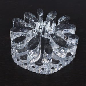 Könnte beinhalten: Ein klarer Glaskerzenhalter mit kreisförmiger Basis und zahlreichen vertikalen, abgerundeten Glaselementen. Das Glas hat ein strukturiertes Aussehen und das Design ist symmetrisch. Der Kerzenhalter ist für mehrere Kerzen konzipiert.