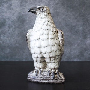 Może przedstawiać: Ceramiczna figurka orła w kolorze białym z brązowymi akcentami. Orzeł ma szczegółową, fakturowaną powierzchnię i stoi na podstawie.