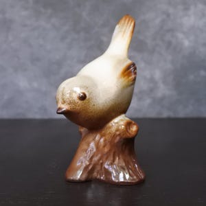 Op de afbeelding: Een keramieken vogelbeeldje in bruin- en crèmetinten. De vogel zit op een bruine boomstronk. De vogel heeft een kleine snavel en een donker oog. Het beeldje is een decoratief item.
