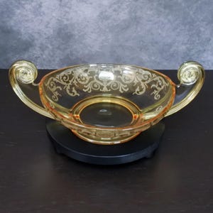 May include: A translucent amber-colored glass bowl featuring an etched floral scrollwork pattern along the rim. The bowl has two prominent, upward-curving scroll handles on opposite sides and rests upon a circular black base.