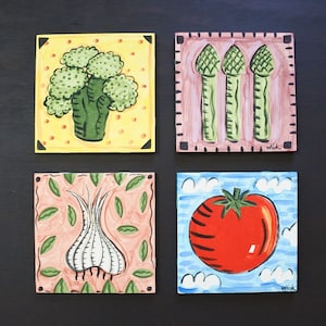 May include: Four square ceramic tiles with hand-painted vegetable designs. The tiles feature a broccoli, asparagus, garlic, and tomato. Each tile has a unique background color and design. The artist's signature, "Wick", is visible on two tiles.