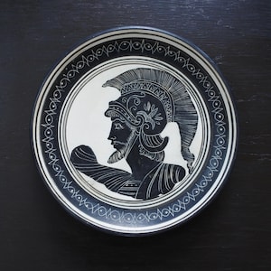 May include: A black and white decorative plate featuring a profile of a warrior in a helmet. The plate has a white center with a black border and a decorative pattern around the edge. The warrior has a beard and is wearing a detailed helmet.