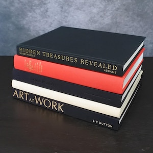 May include: A stack of four hardcover books. The top book is black with gold text reading Hidden Treasures Revealed. Below it is a red book, a black book with embossed text, and a white book with the black text Art at Work and E.P. Dutton.