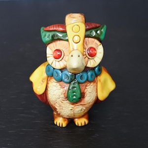 Puede incluir: Figura de búho de cerámica con un diseño detallado. El búho presenta ojos rojos, un pico beige y un tocado decorativo con detalles verdes y amarillos. Tiene un collar de cuentas azul y un colgante verde.