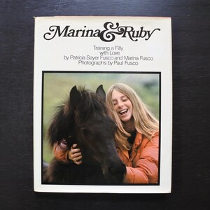 May include: A book cover titled "Marina & Ruby" with the subtitle "Training a Filly with Love." The cover features a photograph of a woman smiling and hugging a black horse. The book's title is in a decorative font.