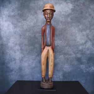 Op de afbeelding: Een gesneden houten beeld van een persoon met een hoed, jas en broek. Het beeld is beschilderd met bruin, rood, blauw en beige. De figuur heeft een slank postuur en staat op een ronde basis.