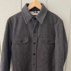 May include: A charcoal gray wool shirt jacket with two chest pockets and a button-down front. The jacket has a collar and long sleeves with buttoned cuffs. The interior lining is a deep burgundy color. The jacket is hanging on a wooden hanger.