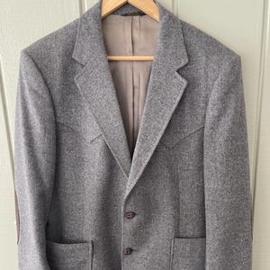 May include: A grey blazer with a two-button fastening, patch pockets, and brown elbow patches. The jacket is suspended on a wooden hanger. The blazer has a notched lapel and three buttons on each cuff. The background is a light-coloured wall.