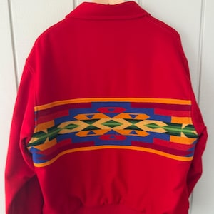 May include: A bright red jacket featuring a horizontal band across the back with a geometric pattern in yellow, blue, green, and dark red. The jacket has a collared neckline, long sleeves, and a ribbed hem, displayed on a wooden hanger.