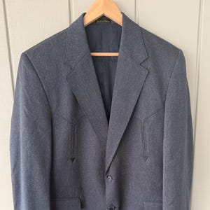 May include: A gray blazer with a classic design, featuring a notched lapel, two front pockets, and decorative stitching on the chest. The blazer is buttoned and hangs on a wooden hanger.