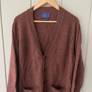 May include: A maroon cardigan sweater with a V-neck, two front pockets, and button closures. The sweater is made of a soft, textured material and is hanging on a wooden hanger. A label is visible on the inside of the sweater.