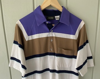 May include: A striped polo shirt with a purple, brown, white, and navy blue color scheme. The shirt has a button-up collar and a pocket on the left side. The shirt has the Jantzen brand name on the left side of the collar.