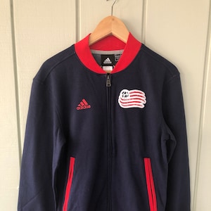 Peut inclure: Une veste bomber bleu marine zippée avec des bordures rouges. La veste porte le logo Adidas sur la poitrine gauche et un patch rouge, blanc et bleu avec un ballon de football sur la poitrine droite. La veste porte également le numéro 1996 sur la manche gauche.
