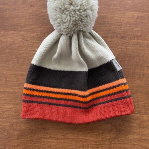Könnte beinhalten: Eine beige Strickmütze mit einem braunen und orangefarbenen Streifenband und einem weißen Bommel oben. Die Mütze hat ein kleines weißes Etikett mit dem Wort "Nordic" darauf.