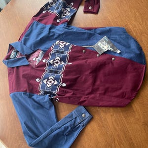 May include: A long-sleeved button-down shirt with a geometric pattern in shades of blue, white, and maroon. The shirt has a pointed collar and a single chest pocket. The label reads "John Deere".