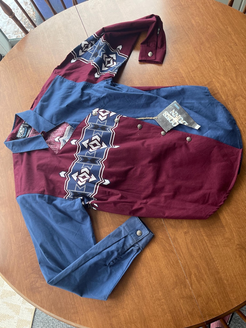 May include: A long-sleeved button-down shirt with a geometric pattern in shades of blue, white, and maroon. The shirt has a pointed collar and a single chest pocket. The label reads "John Deere".