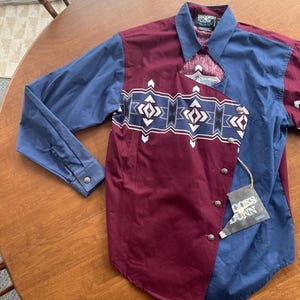 May include: A long-sleeved button-down shirt with a geometric pattern in shades of blue, white, and black. The shirt is made of a burgundy fabric and has a blue and white pattern on the collar. The shirt has a label that says "RODEO  EDDY" and "COORS  LIGHT".