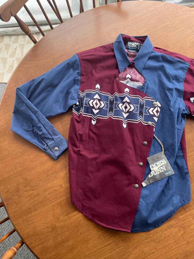 May include: A long-sleeved button-down shirt with a geometric pattern in shades of blue, white, and black. The shirt is made of a burgundy fabric and has a blue and white pattern on the collar. The shirt has a label that says "RODEO  EDDY" and "COORS  LIGHT".