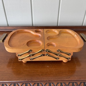 Puede incluir: Una bandeja de servicio de madera vintage con un diseño de bisagra. La bandeja es de color marrón claro con asas y bisagras de color latón. Cuenta con compartimentos tallados para guardar objetos. La bandeja está sobre una superficie de madera oscura.