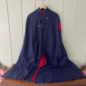 May include: A navy blue cape with a red lining and a high collar. The cape features two sets of button closures and a red emblem on the right shoulder. It is suspended from a wooden hanger.