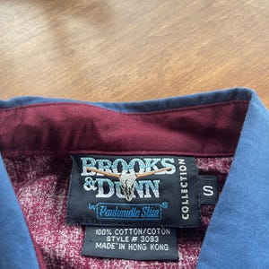 May include: A burgundy and blue button-down shirt with a Brooks & Dunn label. The label features a steer skull logo and the text "Panhandle Slim" in a white circle. The label also includes the text "100% Cotton/Coton, Style # 3093, Made in Hong Kong, Collection, S".