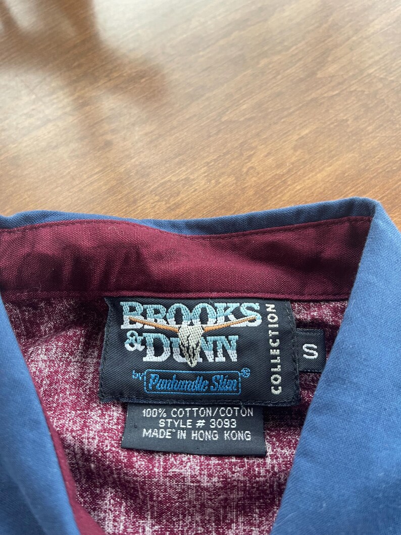 May include: A burgundy and blue button-down shirt with a Brooks & Dunn label. The label features a steer skull logo and the text "Panhandle Slim" in a white circle. The label also includes the text "100% Cotton/Coton, Style # 3093, Made in Hong Kong, Collection, S".