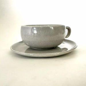 May include: A gray ceramic teacup and saucer set. The cup has a rounded shape and a small handle. The saucer is a simple, round shape.