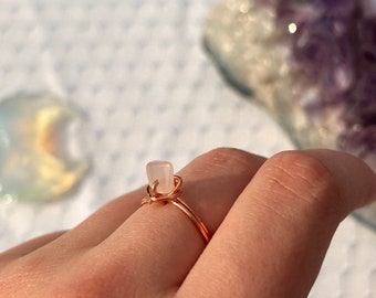 May include: A delicate rose gold ring with a small, white, rectangular gemstone set in the center.