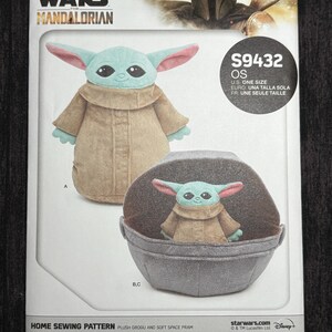 Puede incluir: Un patr&oacute;n de costura Simplicity para un Grogu de peluche y un cochecito espacial suave. El patr&oacute;n muestra el peluche de Grogu con una t&uacute;nica marr&oacute;n con orejas grandes y un cochecito gris. El logotipo de Star Wars Mandalorian est&aacute; en la parte superior.