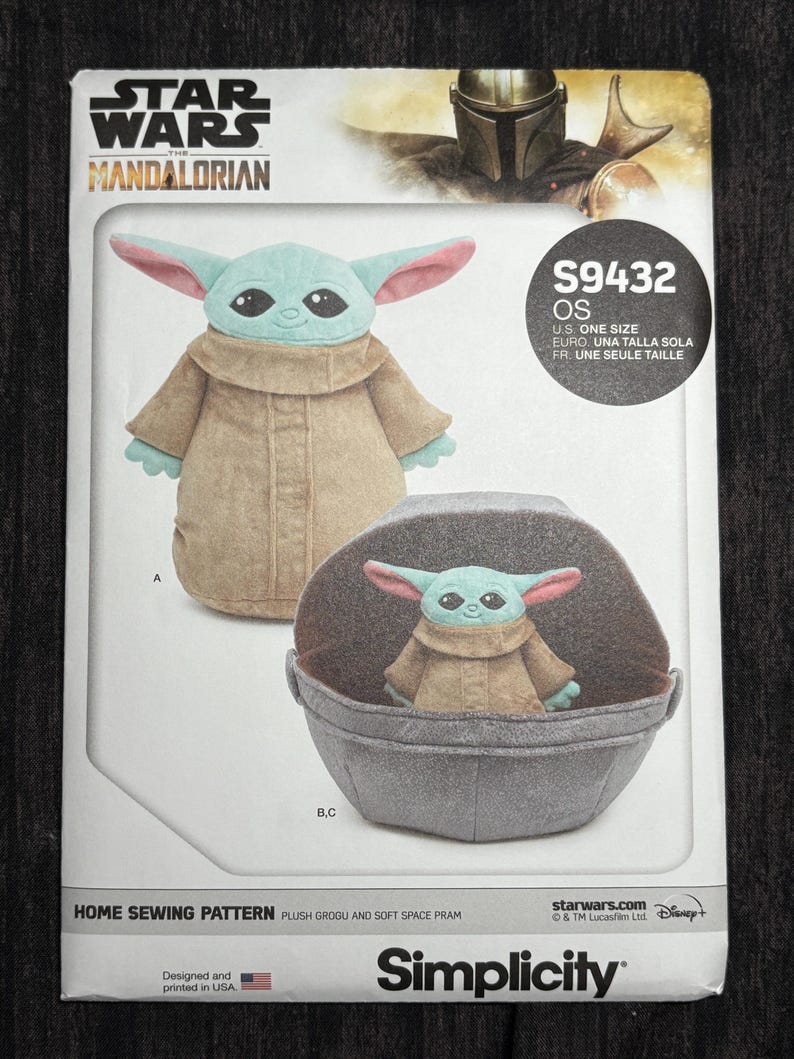 Puede incluir: Un patr&oacute;n de costura Simplicity para un Grogu de peluche y un cochecito espacial suave. El patr&oacute;n muestra el peluche de Grogu con una t&uacute;nica marr&oacute;n con orejas grandes y un cochecito gris. El logotipo de Star Wars Mandalorian est&aacute; en la parte superior.