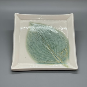 May include: Square ceramic dish with a white border and a large, textured green leaf design in the center. The leaf has subtle yellow and blue-green hues, creating a natural, organic aesthetic. The dish has slightly curved edges.