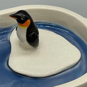 May include: A ceramic penguin figurine with black, white, and orange coloring, perched on a white, cracked-texture iceberg. The iceberg sits in a blue, water-like basin with a white rim. The penguin has a yellow accent.