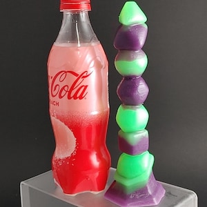 Può includere: Una candela di vetro verde e viola con un design impilato. La candela è appoggiata su una piattaforma in acrilico trasparente accanto a una bottiglia di plastica rossa con la scritta "Coca-Cola" stampata su di essa.