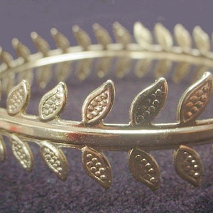 May include: A gold-colored metal headband with a laurel leaf design. The leaves are evenly spaced around the band and have a textured surface. The headband is likely for decorative purposes.