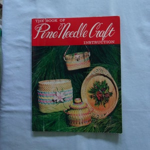 May include: A vintage book titled "The Book of Pine Needle Craft Instruction" with a red cover. The cover features images of woven pine needle crafts, including baskets and a decorative tray, set against a backdrop of green pine needles.