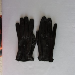 May include: A pair of black leather gloves with zippers on the back of the wrists. The gloves are lying flat on a white surface, showcasing their sleek design and detailing. The gloves appear to be new.