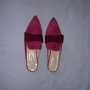 May include: A pair of fuchsia-colored slip-on shoes with pointed toes and a dark burgundy bow detail. The shoes have a suede-like texture and a neutral-toned insole. The footwear is presented on a plain, light-colored surface.