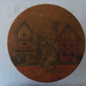 May include: A round, wooden decorative box with a painted cat and birdhouse scene. The cat is brown and tan, with a red collar. The birdhouse and house are red and brown, with a green bird flying above.