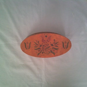 May include: Oval-shaped, orange wooden box with a floral design. The box features a dark floral pattern in the center and on each end. The box is likely used for storage or decoration.