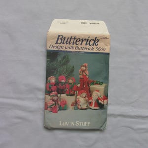 May include: A vintage Butterick sewing pattern envelope, featuring a colorful illustration of Christmas-themed fabric dolls. The envelope reads "Butterick Design with Butterick 5600" and "Luv 'n Stuff". The background is a light gray.