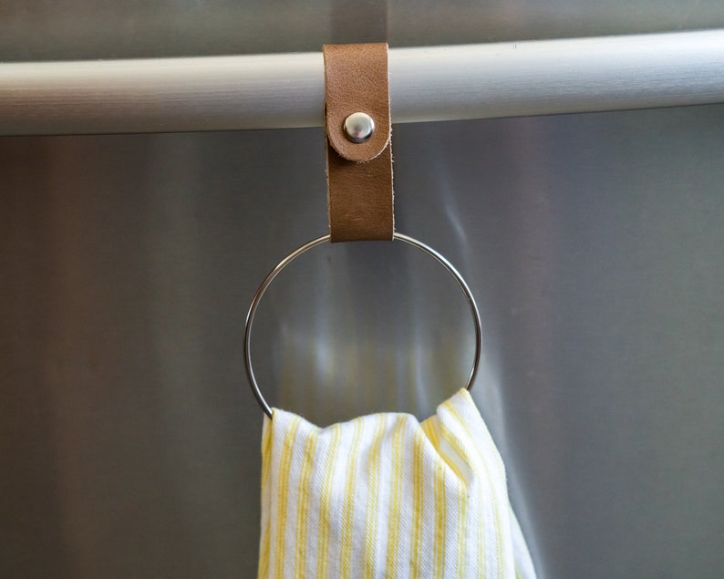 May include: A brown leather strap with a metal ring attached to a stainless steel bar. A yellow and white striped kitchen towel is hanging on the ring.