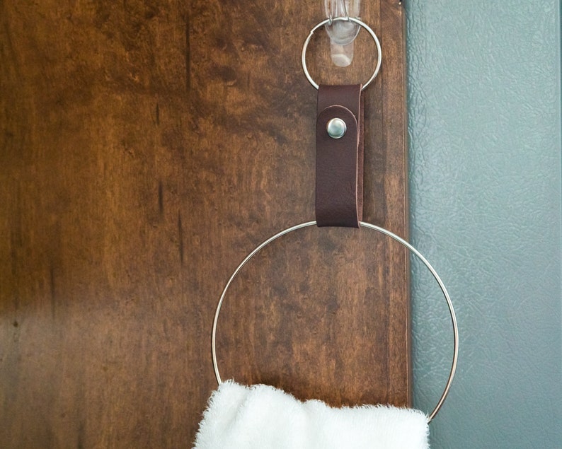 May include: A brown leather strap with a metal ring attached to a silver hook. The ring is holding a white towel.