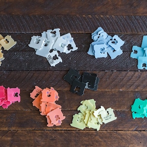 May include: A collection of plastic bread tags in various colors including beige, white, light blue, cyan, pink, orange, black, yellow, and green. Each tag features a central heart-shaped cutout and some display printed text such as dates.